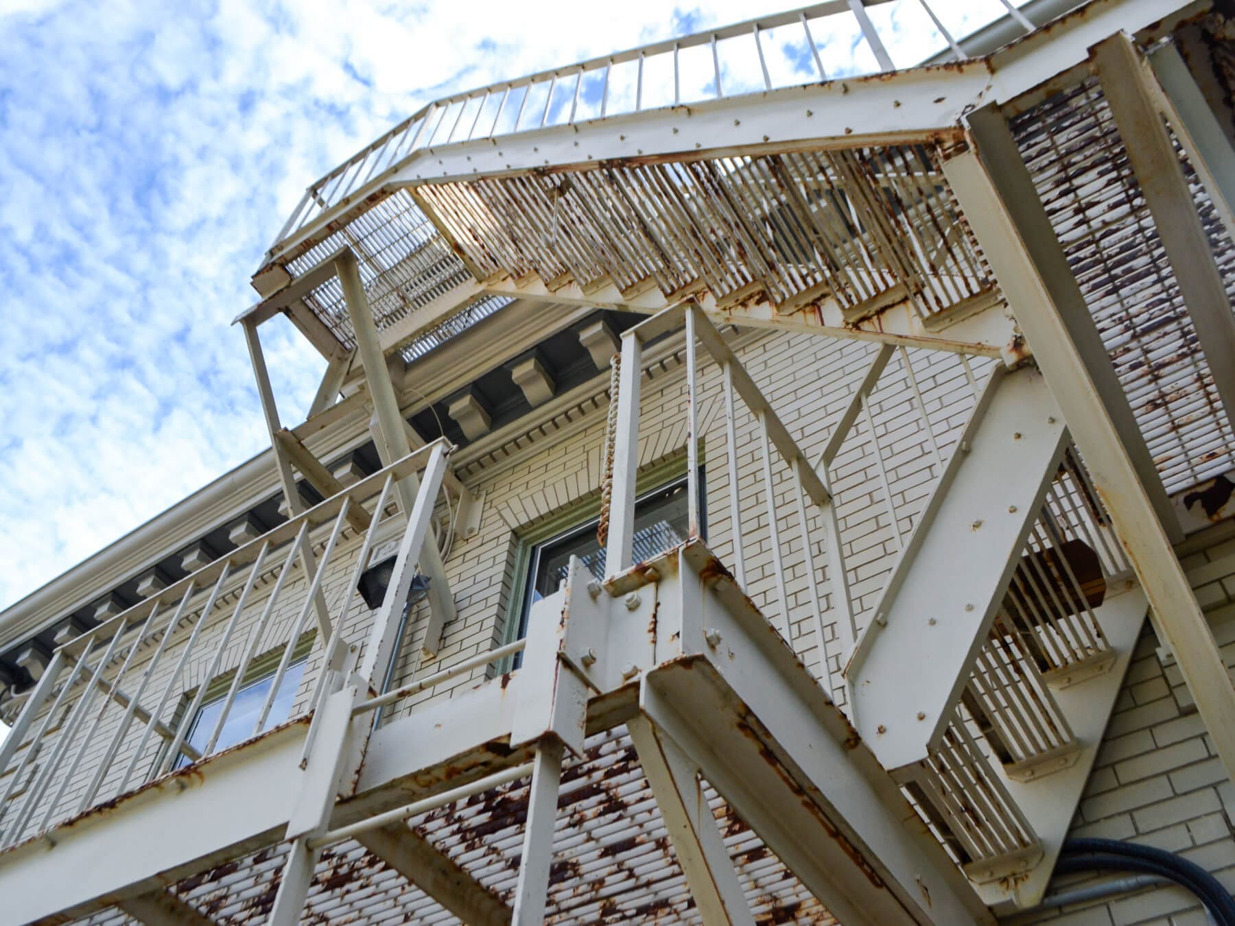 Rusty metal fire escape on a brick building, viewed from below against a blue sky.