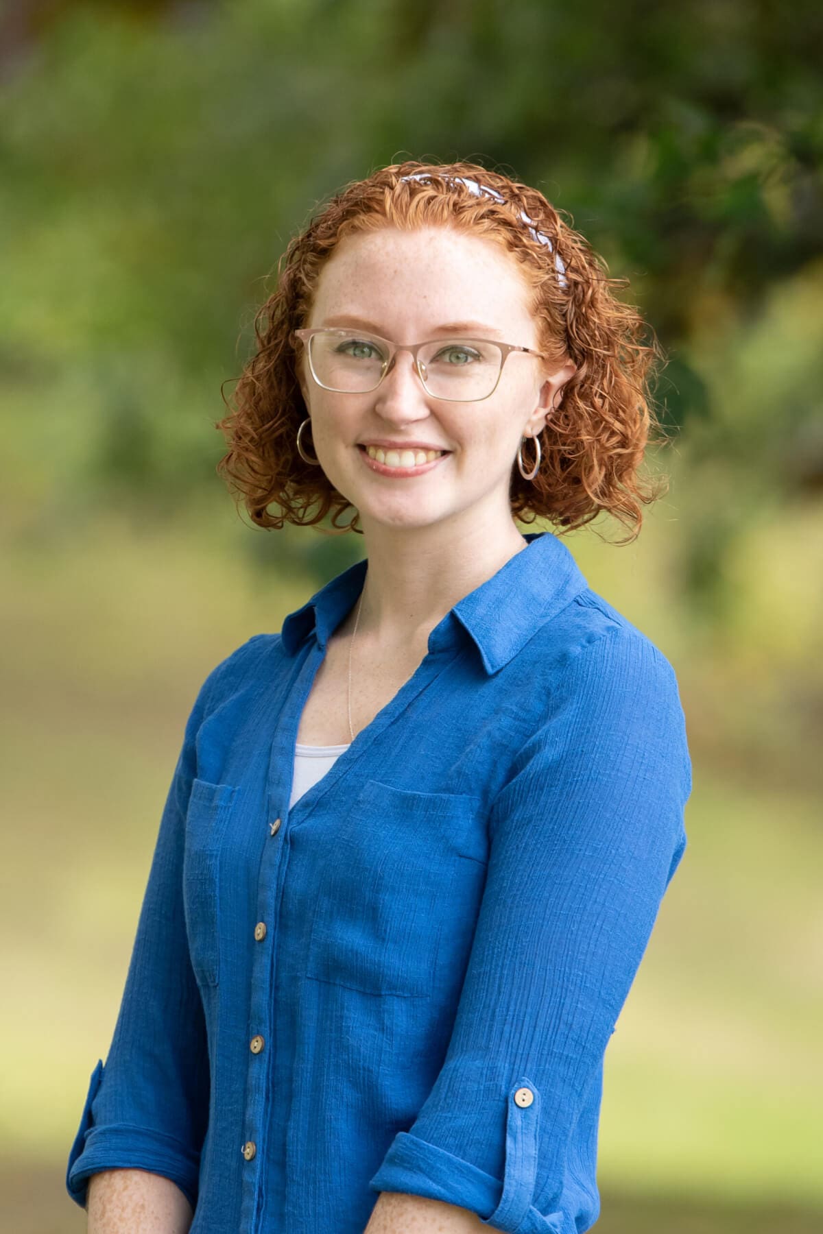 Professional headshot of woman standing outdoors with a blurred green background.