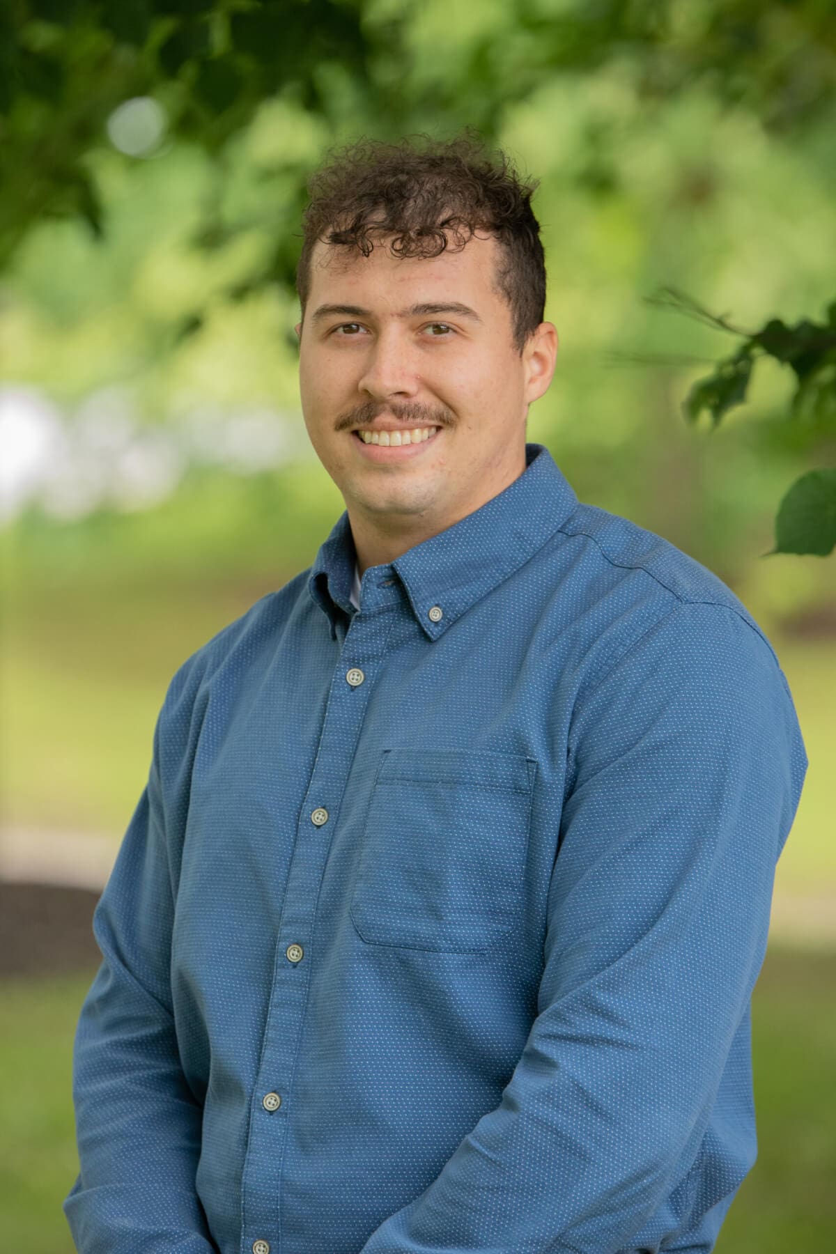 Professional headshot of man standing outdoors with a blurred green background.