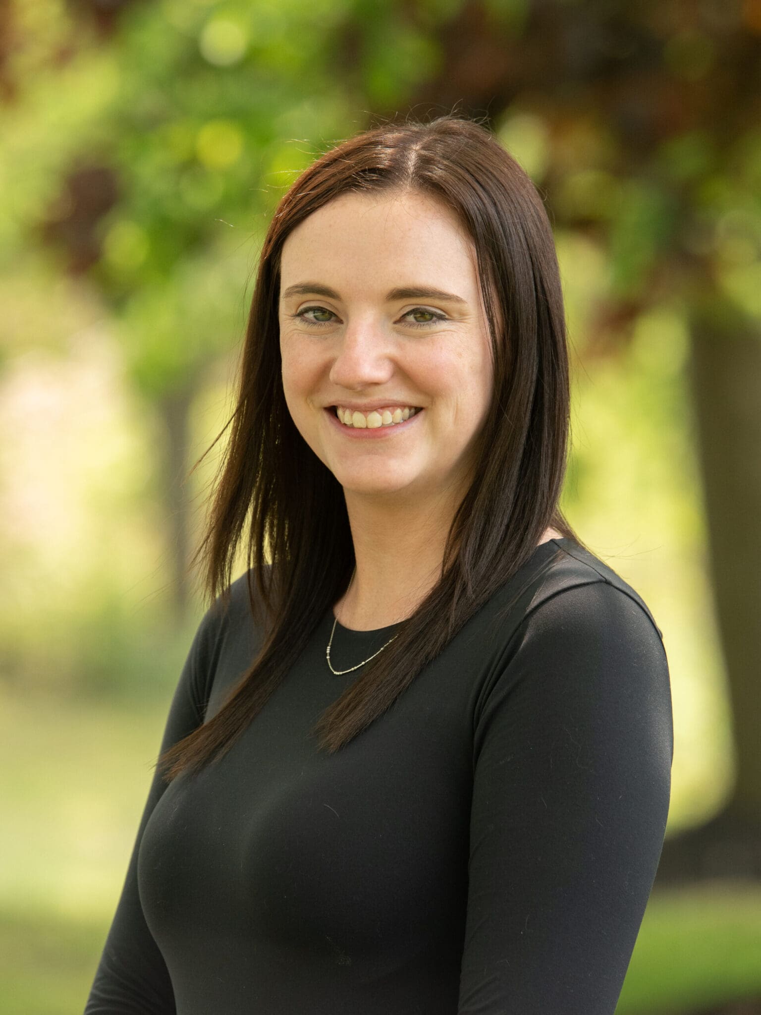 Professional headshot of woman standing outdoors with a blurred green background.