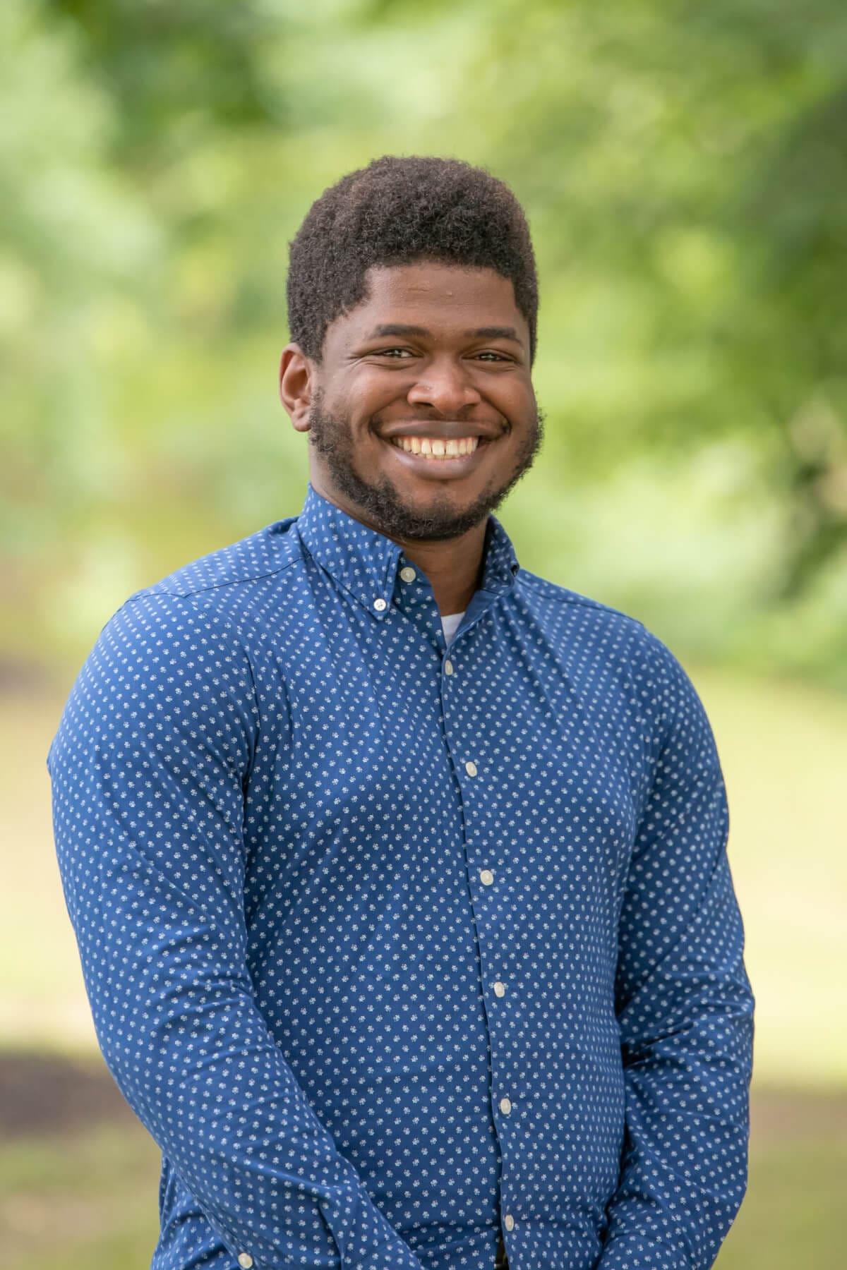 Professional headshot of man standing outdoors with a blurred green background.