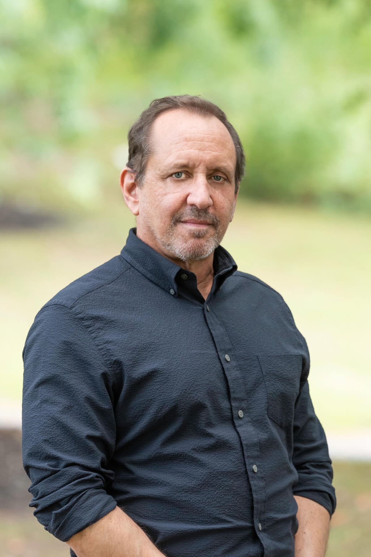 Professional headshot of man standing outdoors with a blurred green background.