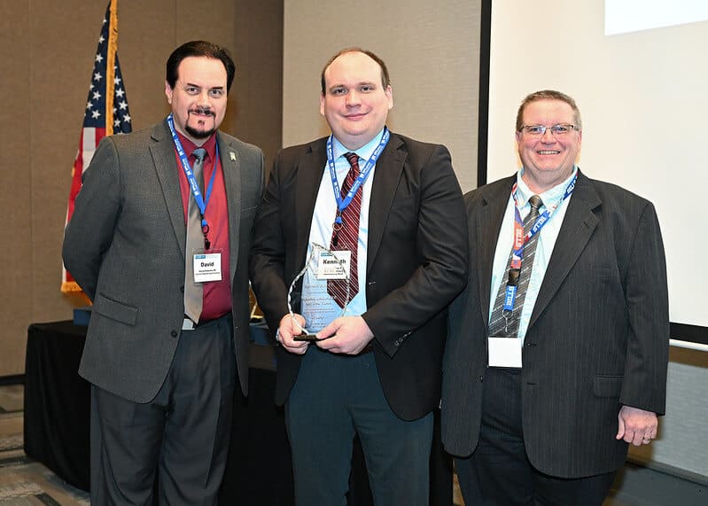 Men in suits at an award ceremony holding a trophy, standing in front of an American flag.