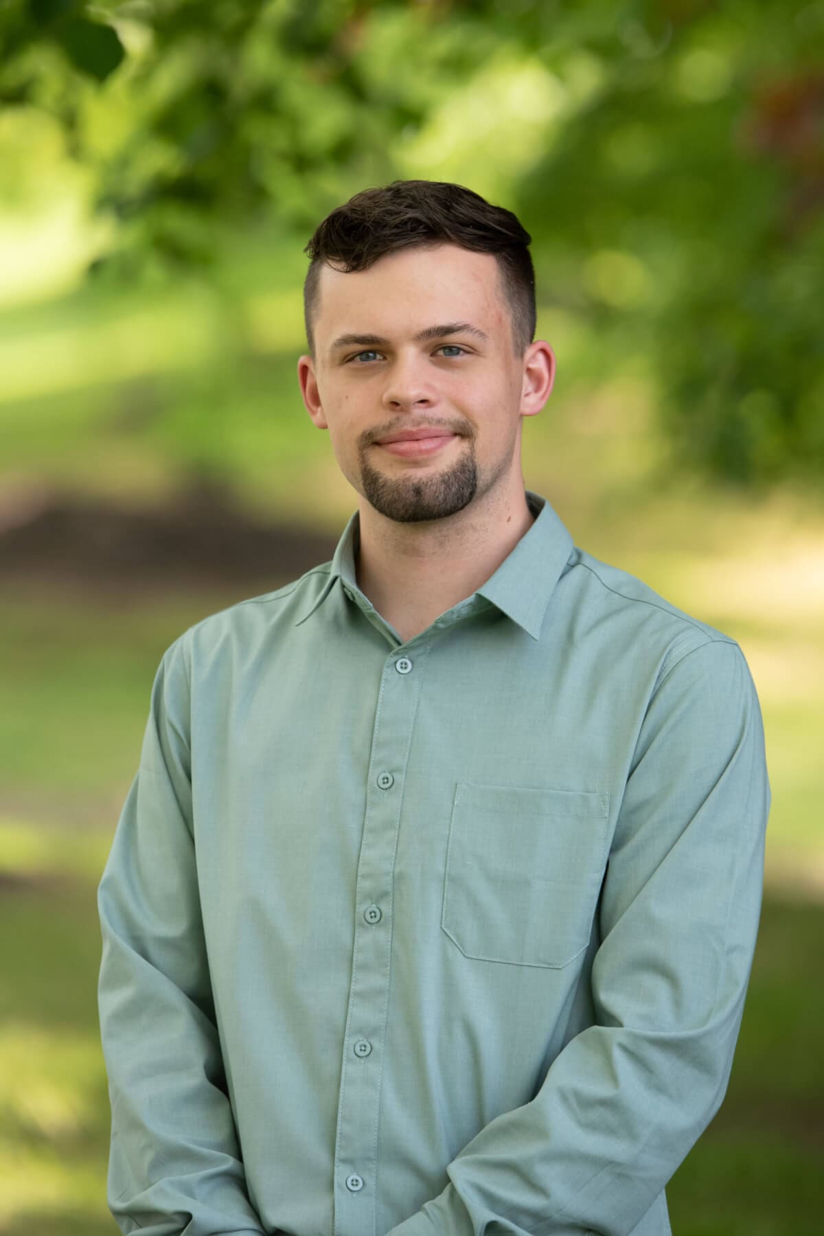 Professional headshot of man standing outdoors with a blurred green background.