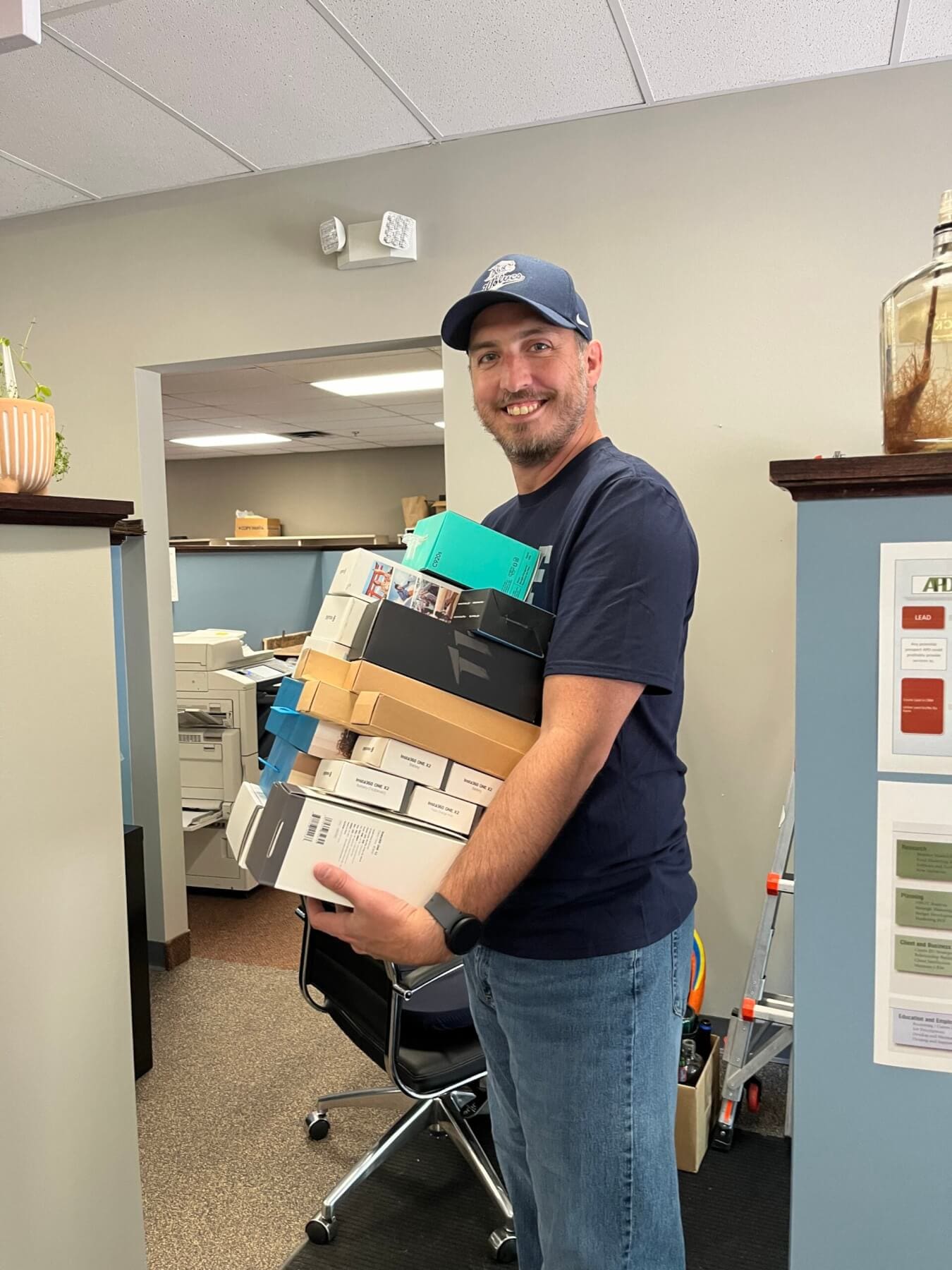 Man carrying multiple office supply boxes in a workspace, smiling at the camera.