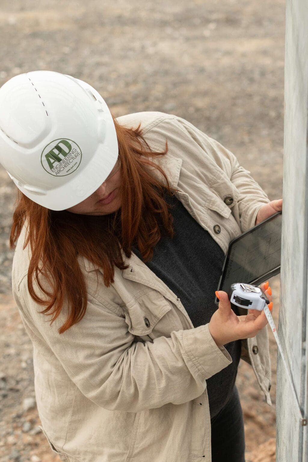 Woman in a hard hat using a tablet and tape measure on a construction site.
