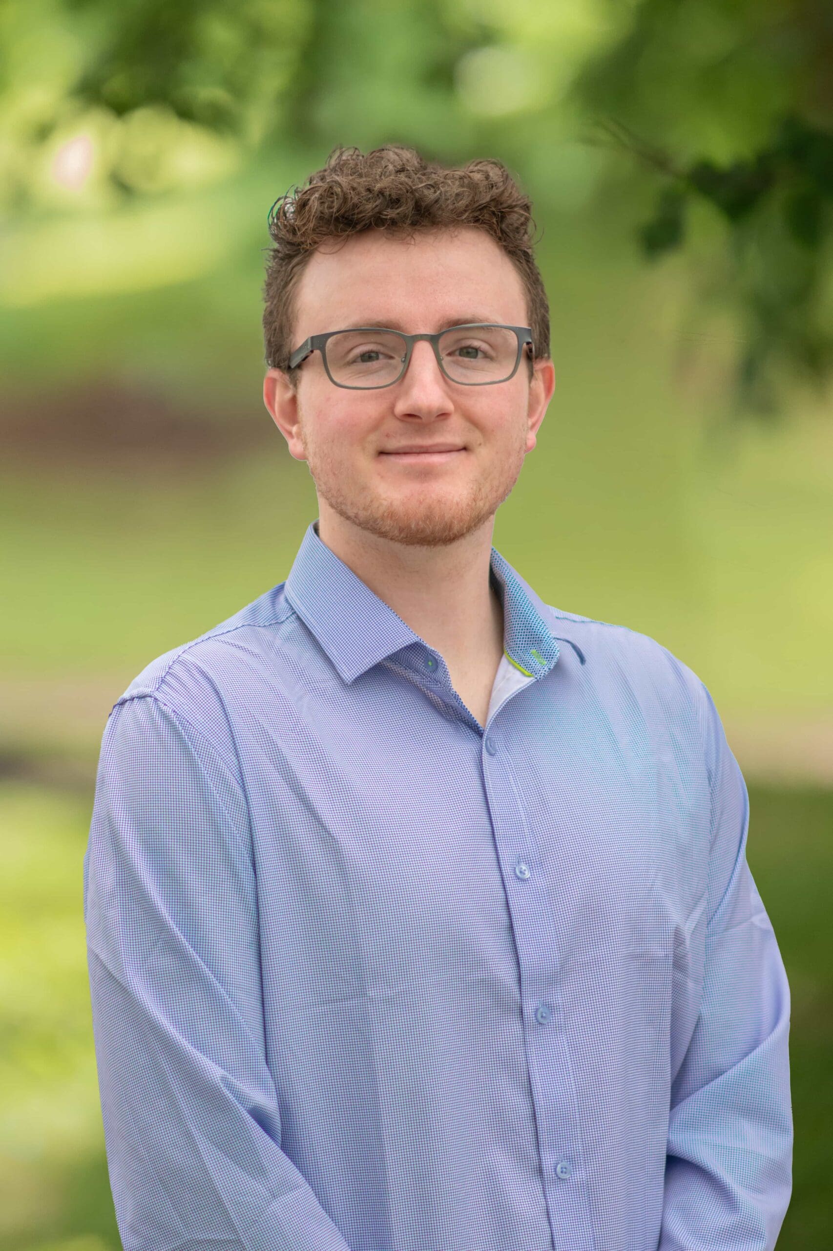 Young professional in a blue shirt, standing outdoors with a blurred green background.