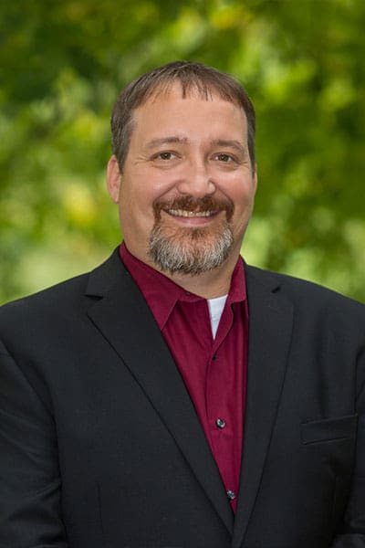 Professional headshot of man standing outdoors with a blurred green background.
