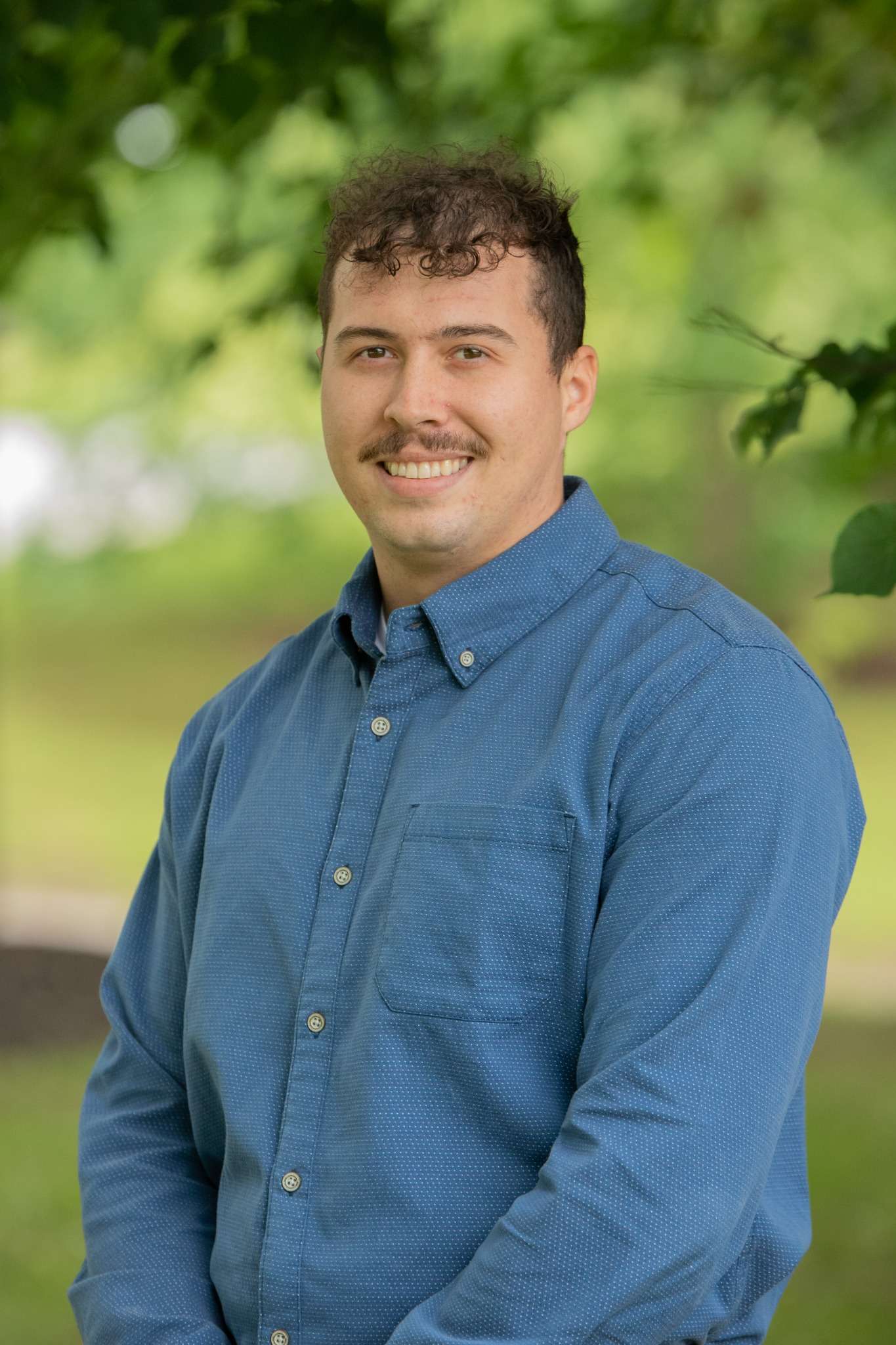Professional headshot of man standing outdoors with a blurred green background.