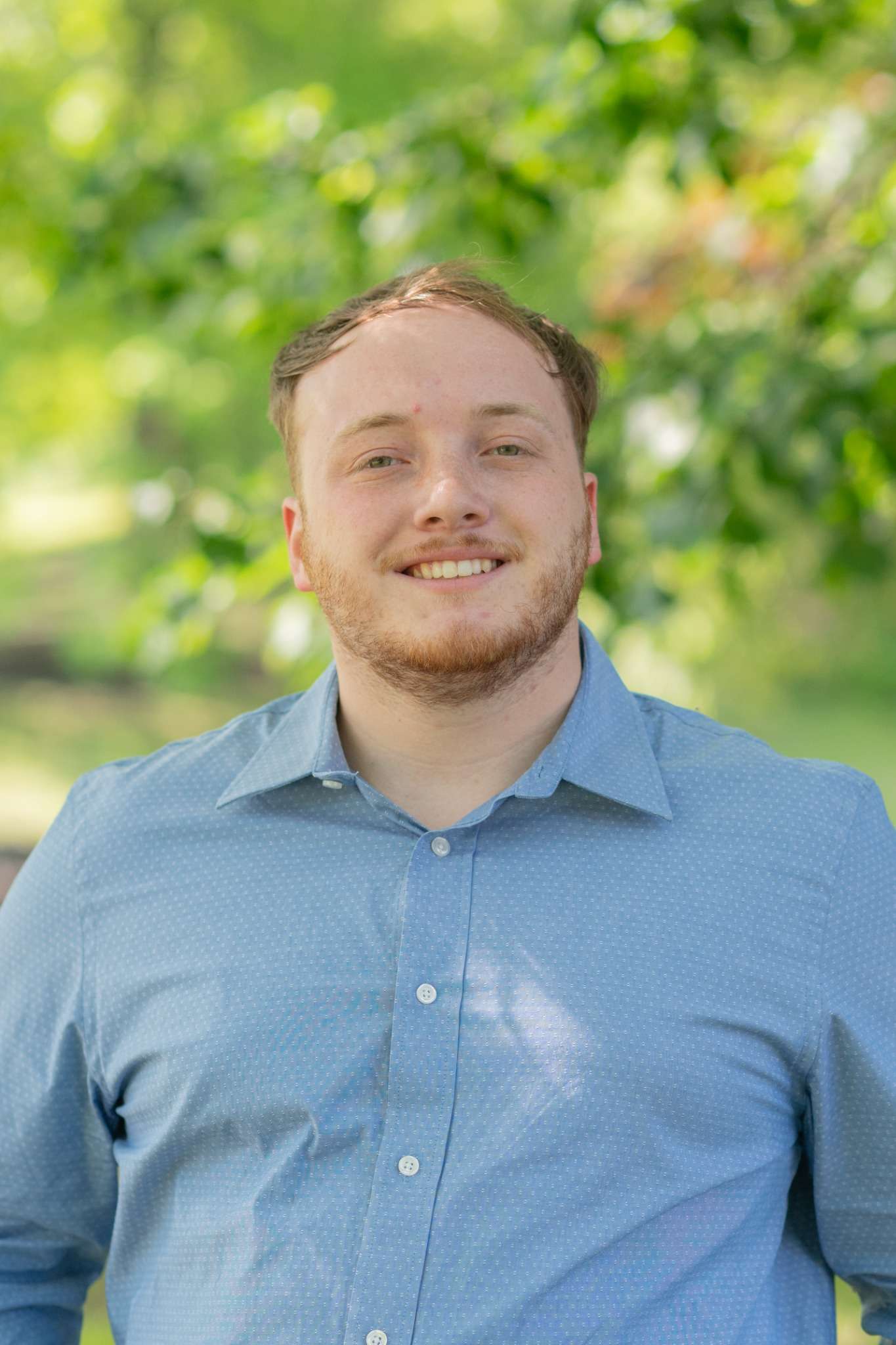 A man in a blue shirt smiling outdoors, standing against a backdrop of lush green trees.
