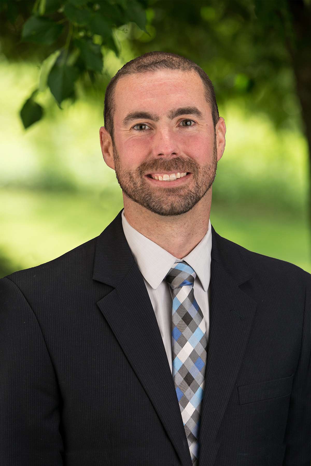 Professional headshot of man standing outdoors with a blurred green background.