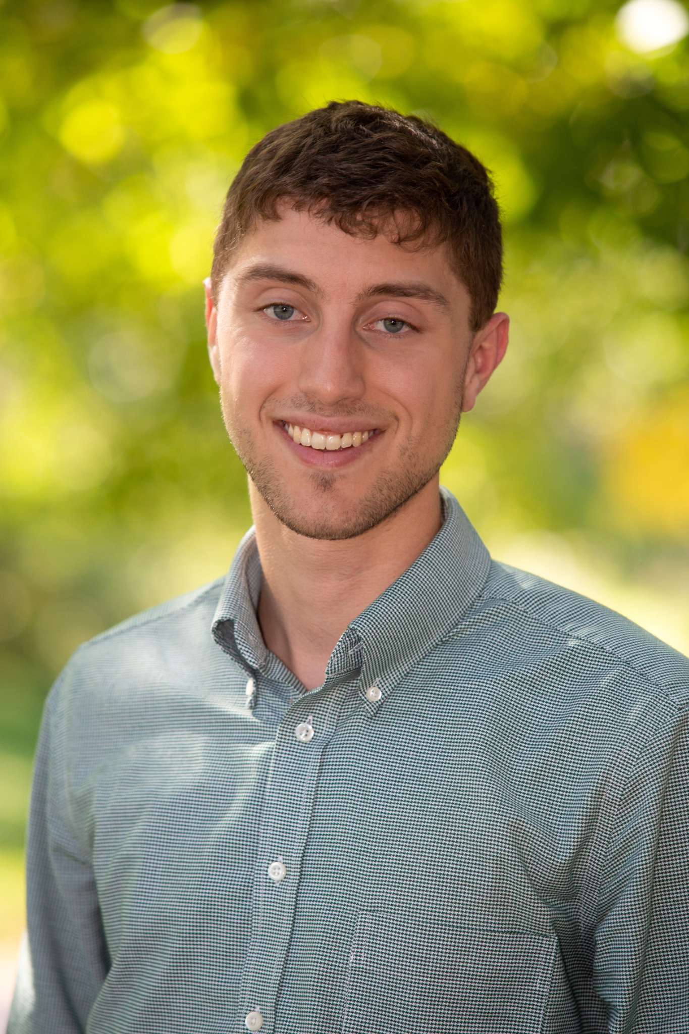 Professional headshot of man standing outdoors with a blurred green background.