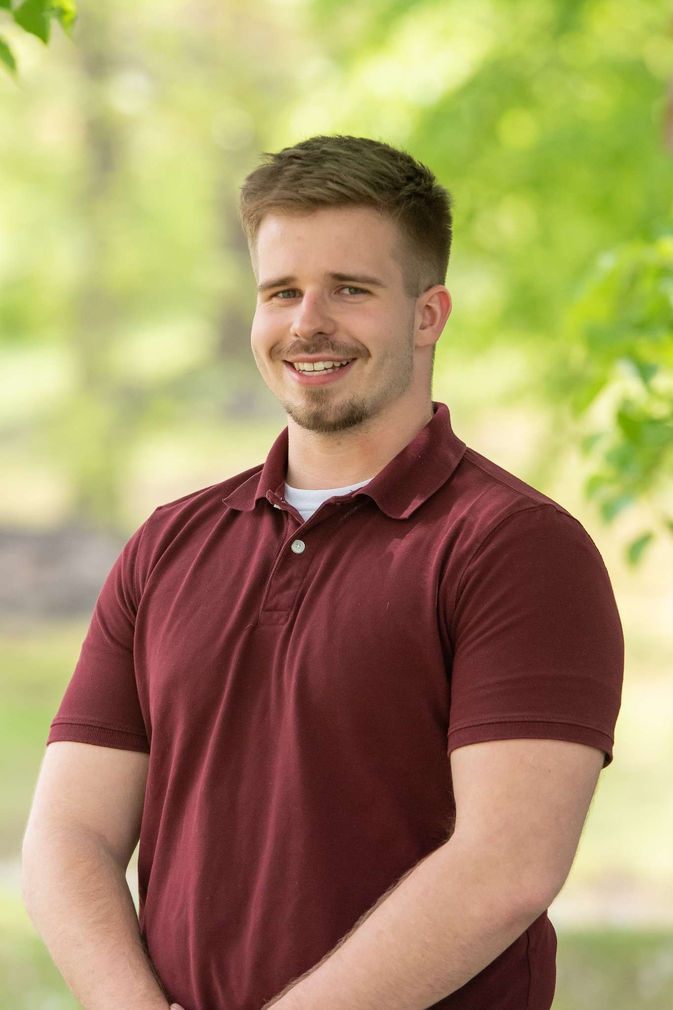 Professional headshot of man standing outdoors with a blurred green background.