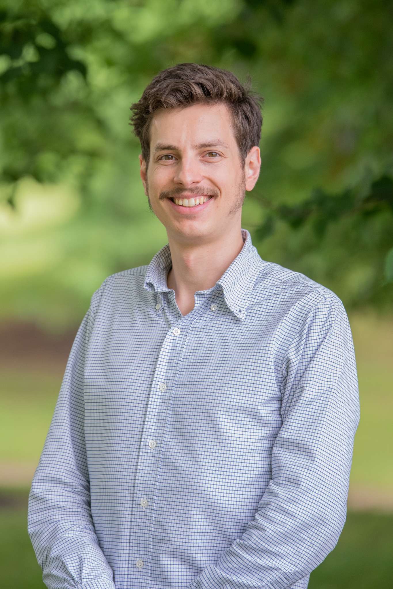 Professional headshot of man standing outdoors with a blurred green background.
