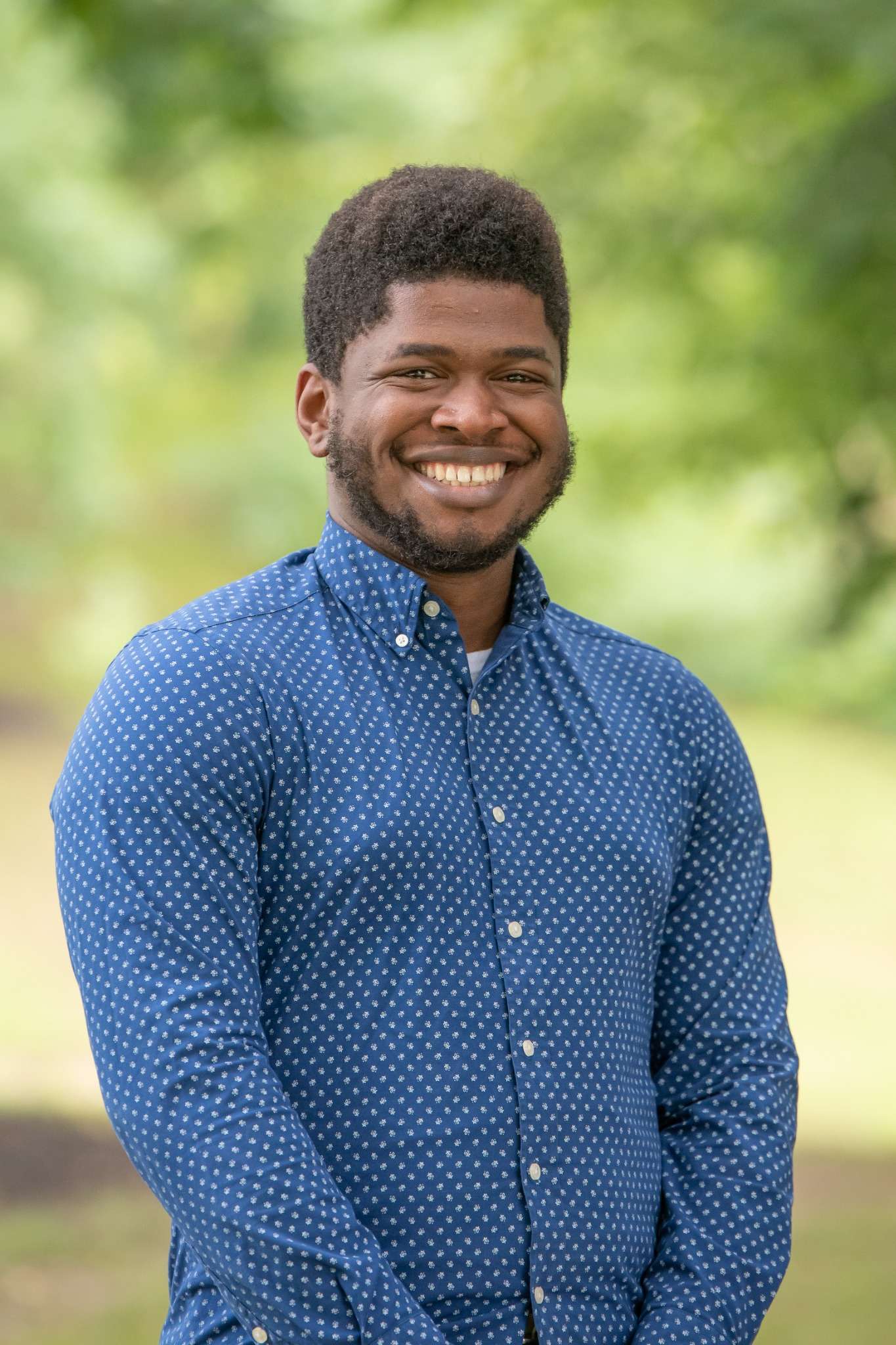 Professional headshot of man standing outdoors with a blurred green background.