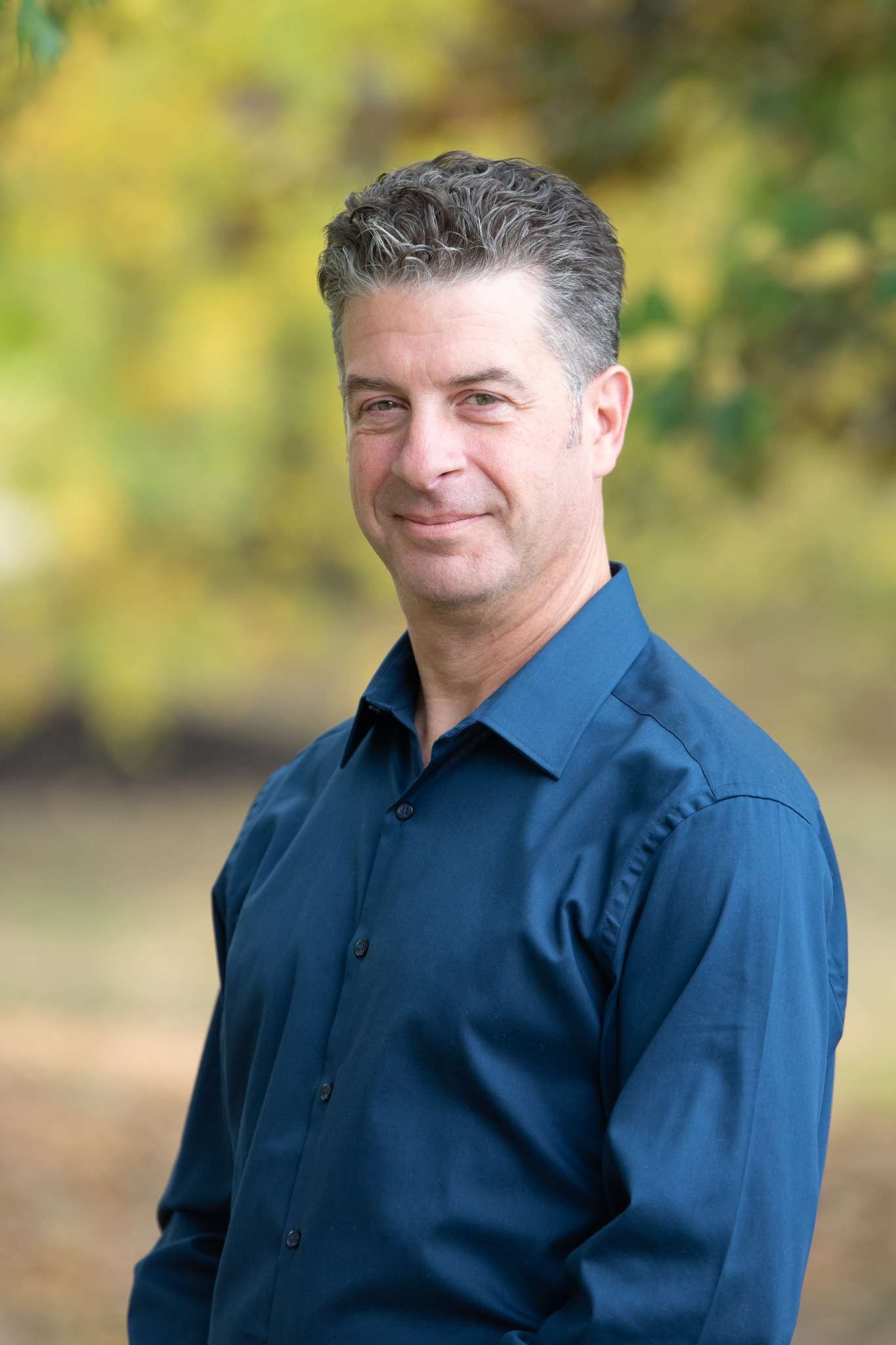Professional headshot of man standing outdoors with a blurred green background.