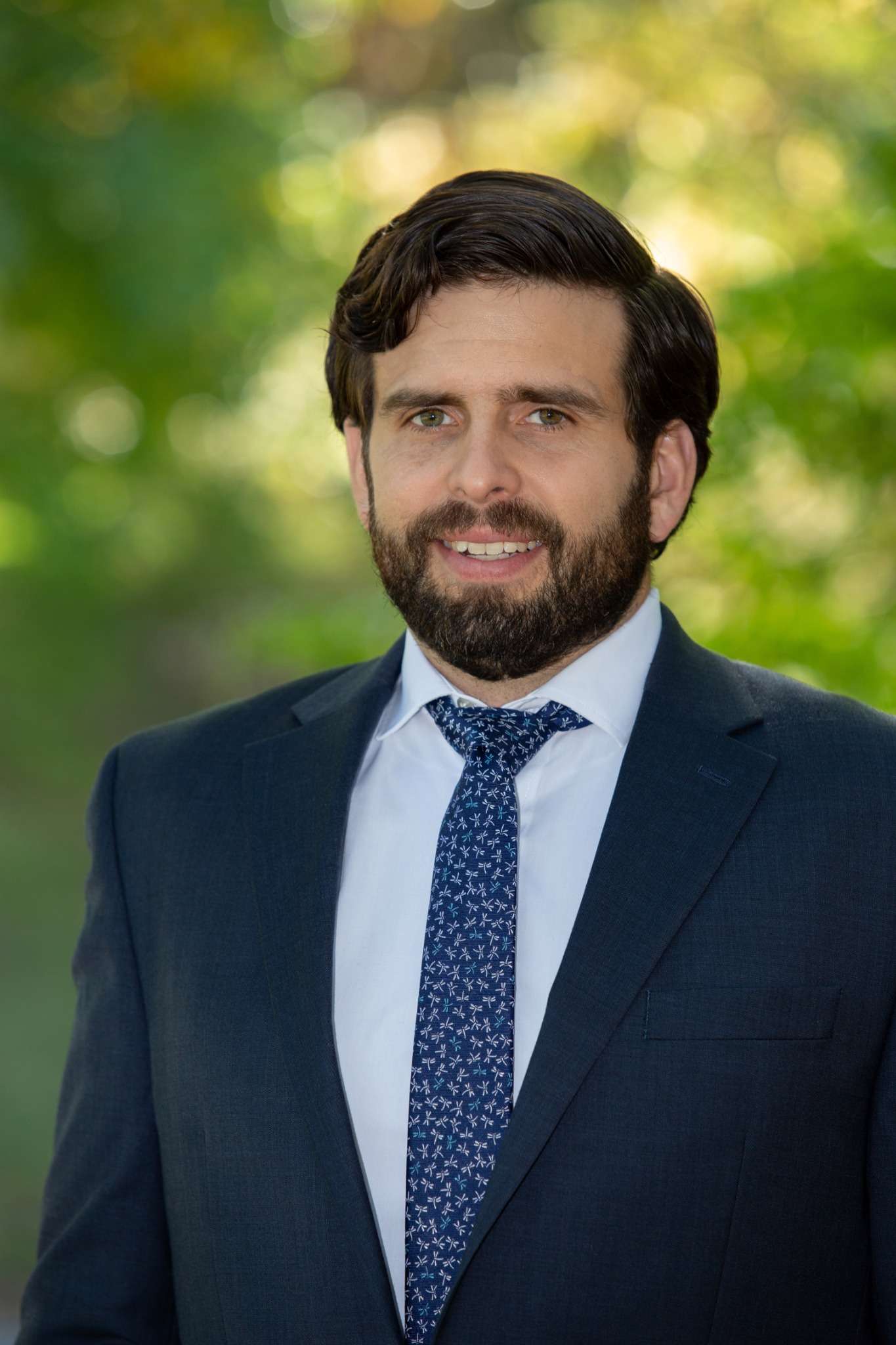 Professional headshot of man standing outdoors with a blurred green background.