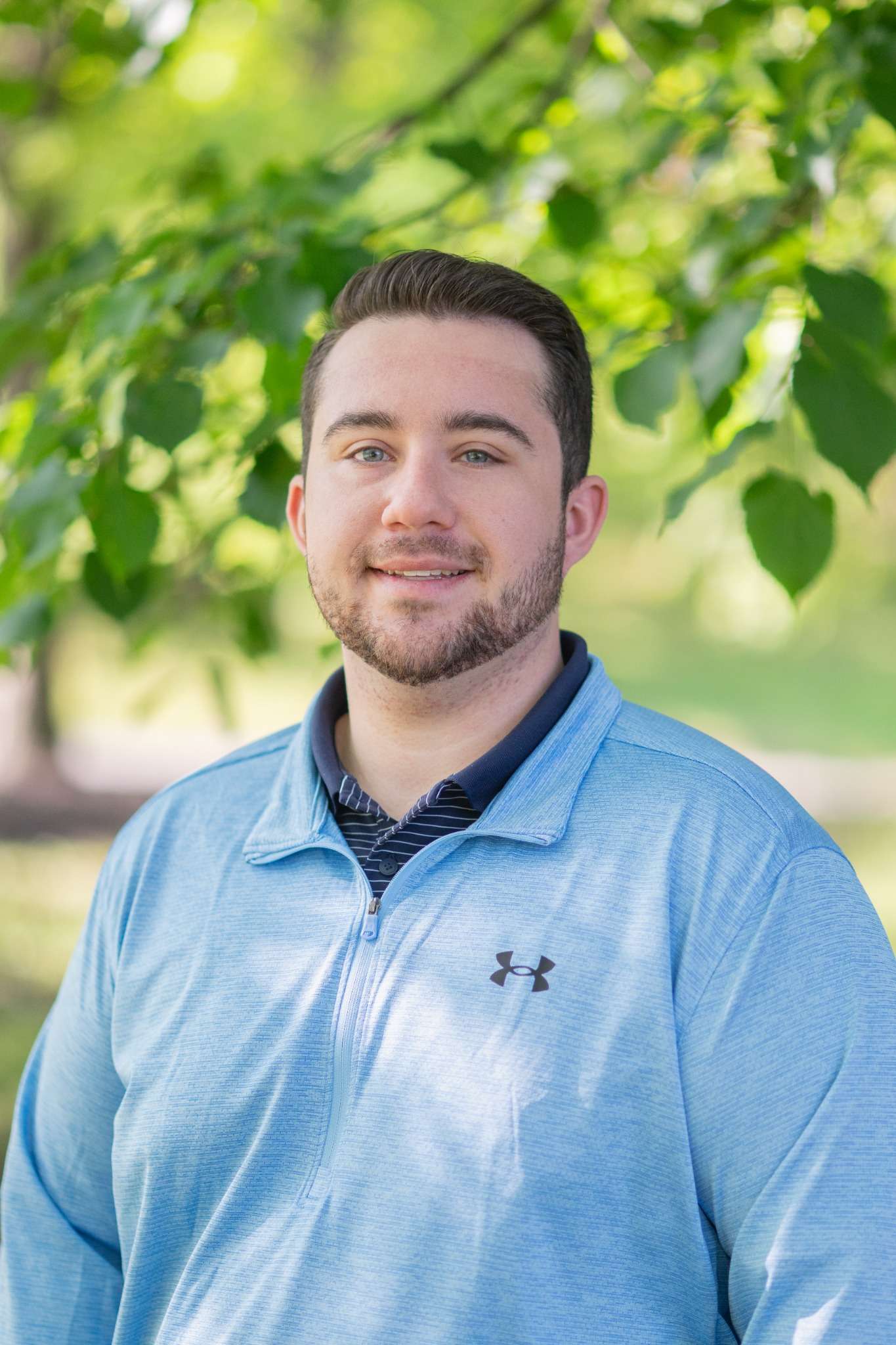 Professional headshot of man standing outdoors with a blurred green background.