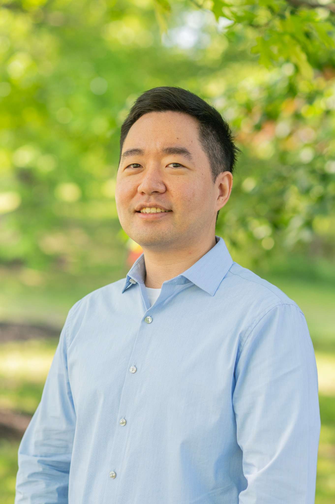 Professional headshot of man standing outdoors with a blurred green background.