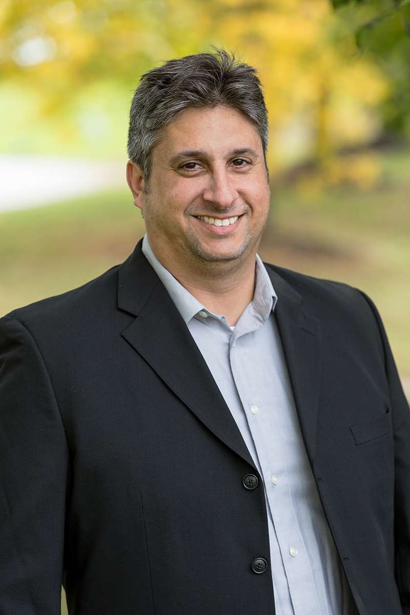 Professional headshot of man standing outdoors with a blurred green background.