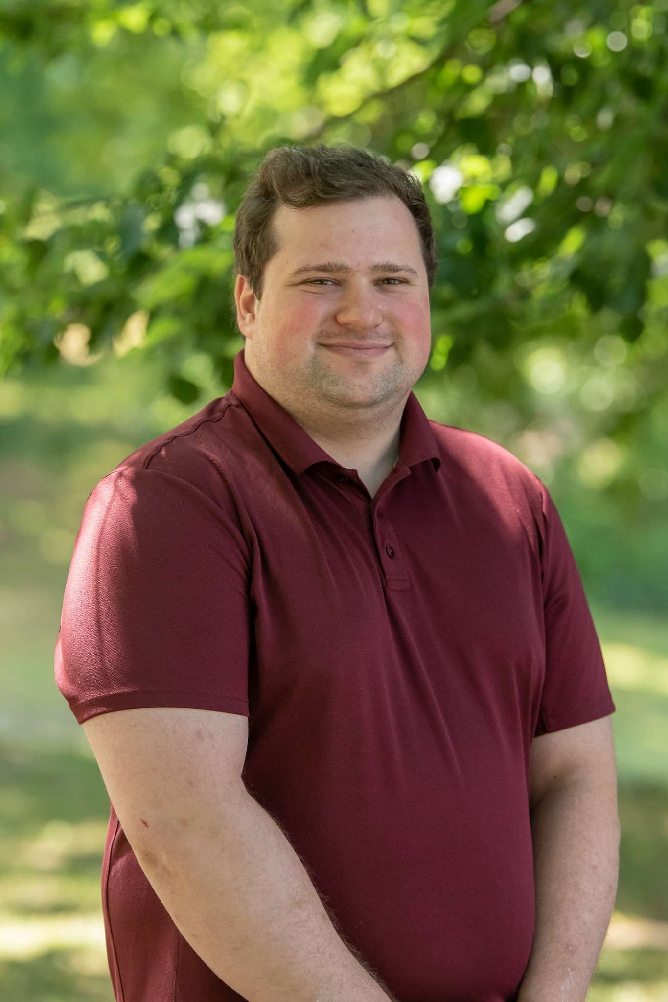 A smiling man in a maroon polo shirt stands outdoors with trees in the background.