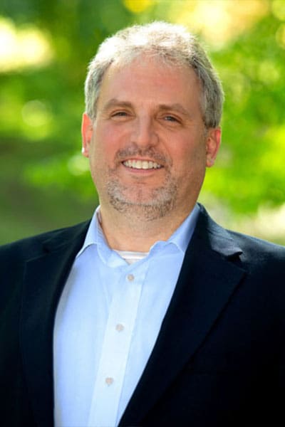 Professional headshot of man standing outdoors with a blurred green background.
