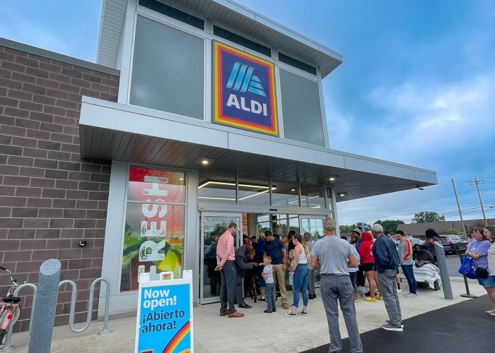 Customers gather at ALDI grand opening with sign reading Now open! ¡Abierto ahora! against a clear blue sky.