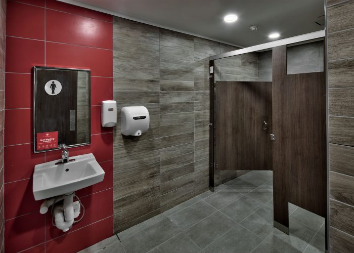 Modern restroom with red and wood paneling, sink, and stalls, featuring hand dryer for hygiene.