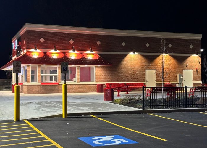 Exterior view of a fast-food restaurant with red accents at night, featuring outdoor seating and accessible parking.