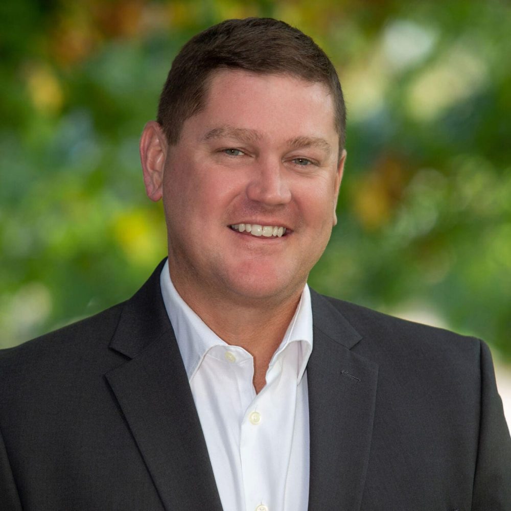Professional headshot of man standing outdoors with a blurred green background.