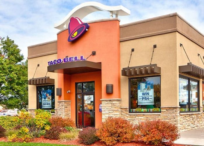 Exterior of a Taco Bell restaurant with a landscaped surrounding, showcasing brand signage under a blue sky.