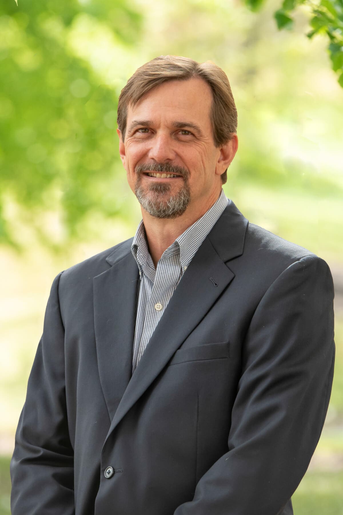 Professional headshot of man standing outdoors with a blurred green background.